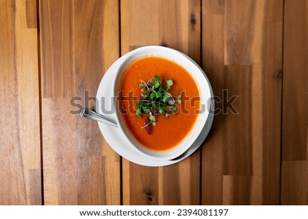 Velvety tomato soup topped with fresh microgreens, served in a white bowl against a warm wooden backdrop, captured from above for a comforting visual feast