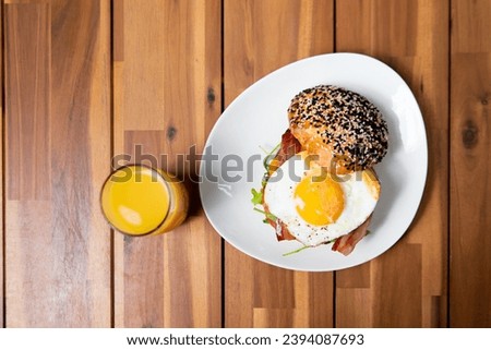 A freshly prepared breakfast sandwich with a multigrain bagel, topped with sesame and poppy seeds, a sunny-side-up egg, bacon, and arugula, served with a glass of orange juice on a wooden table