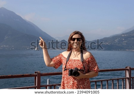 Happy brunette 30s woman enjoying summer holidays. Concept of vacation and travelling. Varenna, Italy. Lake Como. Redhead girl pointing finger and holding a camera outdoors at summer day