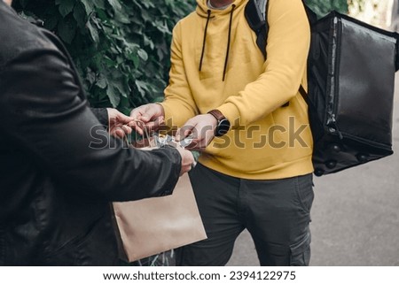 Man pays for food delivery close up .Male courier food delivery man in positive in uniform hands over food order and receives money tips payment from man client in jacket.  Royalty-Free Stock Photo #2394122975