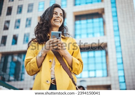 Beautiful happy young woman messaging on smartphone on the background of a city street on a sunny day. Business woman is looking away and smiling. She is standing in the city with a smartphone.
