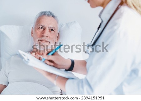 Senior man with female doctor writing to clipboard at hospital ward. Nurse medical worker taking notes of medical history, complaints, prescribing drugs pills treatment