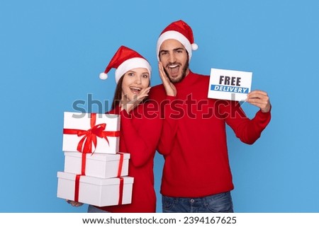 Excited Man And Woman Holding Christmas Gift Boxes And Free Delivery Certificate, Smiling Couple Wearing Santa Hats Enjoying Holiday Shopping And Presents Purchasing, Standing Over Blue Background