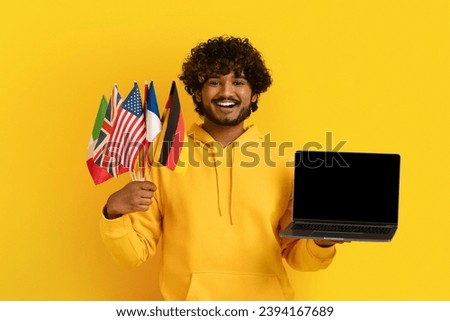 Smiling handsome young indian man wearing yellow hoodie student holding laptop with empty black screen and flags of different countries, yellow background. Education abroad, study online at university