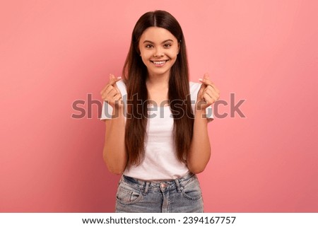 Portrait of lovely smiling teen girl making Korean finger mini heart gesture while standing on pink studio background, beautiful female teenager showing like or love sign, crossing fingers Royalty-Free Stock Photo #2394167757
