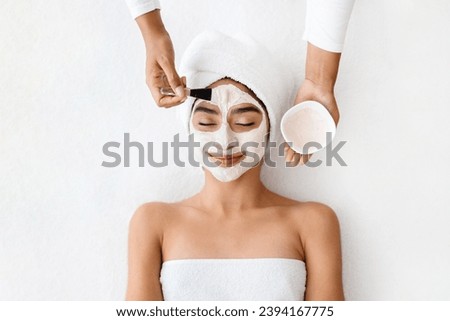 Top view of cosmetologist applying clay mask on face of young indian woman, beautiful eastern lady with towel on head lying on table, enjoying beauty treatments in spa salon, white background Royalty-Free Stock Photo #2394167775