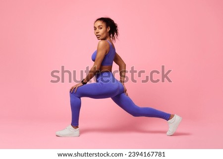 Sporty black woman lunging forward exercising during workout over pink studio background, looking at camera with confidence. Gym training for muscles flexibility. Side view shot