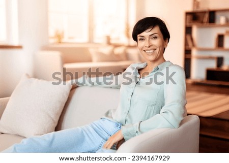 Cheerful middle aged lady smiling looking at camera while sitting elegantly on her living room sofa indoor, embodying relaxed weekend at home. Free space for text Royalty-Free Stock Photo #2394167929