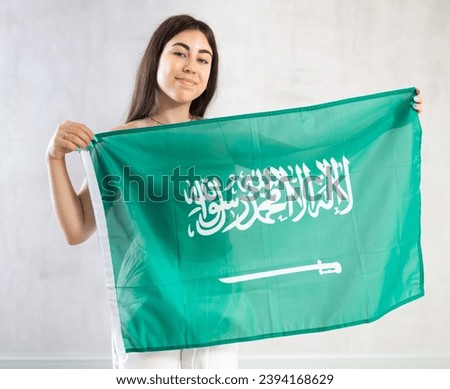 Happy young woman holding large flag of Saudi Arabia posing against background of light unicoloured wall