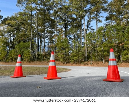 Three orange and white traffic cones blocking off the entrance to a side street. The image is taken from a low angle during the day and there are trees and blue sky. in the background. Royalty-Free Stock Photo #2394172061