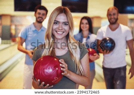 Happy young friends are holding balls, looking at camera and smiling while playing bowling together, girl in the foreground