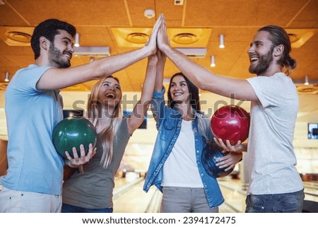 Happy young friends are holding balls, giving high five and smiling while playing bowling together