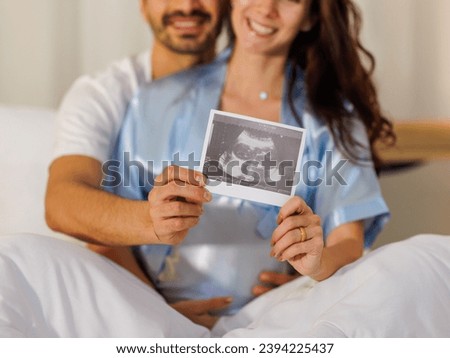 Cheerful husband embracing happy wife on bed at night and showing pregnancy ultrasound image film of unborn baby while mom enjoy holding belly and smile for hopeful of warm family when child birth.