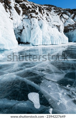 Frozen Baikal Lake on sunny cold winter day. View of ice grotto in coastal rocks on Olkhon Island from ice field with beautiful clear blue ice with cracks. Ice travel and adventure on frozen lake Royalty-Free Stock Photo #2394227649