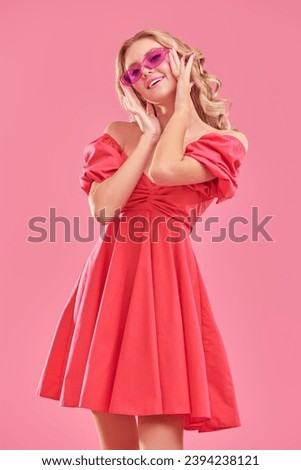 Young beauty. A cute attractive girl with blond hair poses in a pink dress and pink glasses and smiles. Pink studio background. Femininity, beauty and fashion.