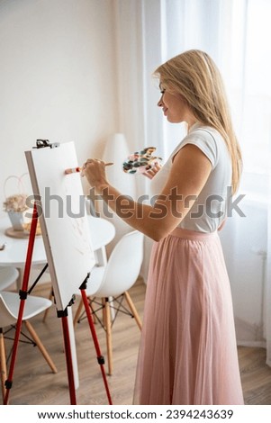 Young woman artist with palette and brush painting abstract white-pink picture on canvas at home. Art and creativity concept. High quality photo