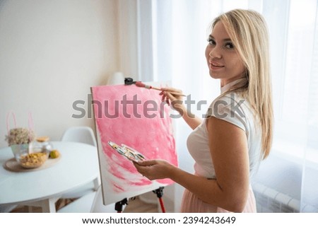 Portrait of young woman artist with palette and brush painting abstract pink picture on canvas at home. Art and creativity concept. High quality photo