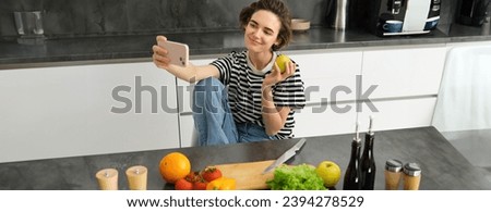 Portrait of smiling brunette woman in the kitchen, posing with an apple, taking selfie on smartphone, cooking and making pictures for internet blog.