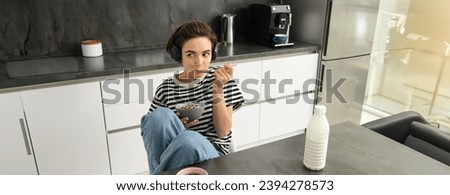 Portrait of smiling young candid woman, student eating morning cereals, having her breakfast, listening music in wireless headphones, sitting in the kitchen. Royalty-Free Stock Photo #2394278573