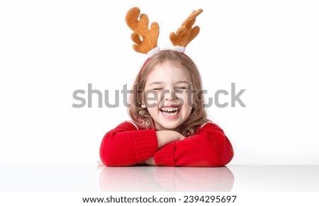 Happy funny caucasian child girl in christmas costume sitting at white empty space table. Holiday advertising blank.