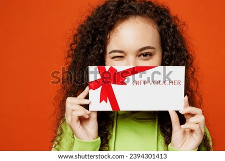 Close young woman of African American ethnicity wearing green hoody casual clothes hold gift certificate coupon voucher card for store wink isolated on plain red orange background. Lifestyle concept