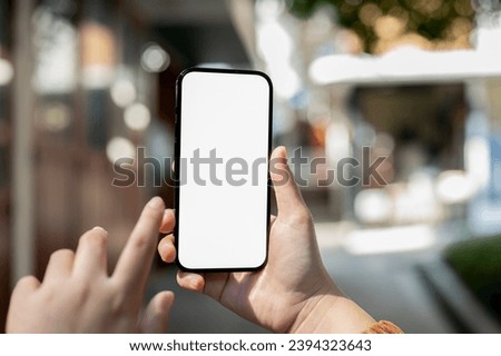 Close-up image of a white-screen smartphone mockup in a woman's hand with a blurred background of retail shops on the street. mobile banking, using map on phone, texting