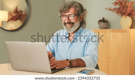 Happy senior man with gray hair wearing glasses, in living-room making online video call using laptop talking Royalty-Free Stock Photo #2394332735