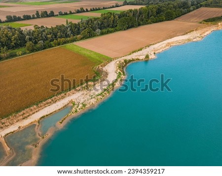 Shore of a quarry pond between fields and trees with turquoise water as aerial view