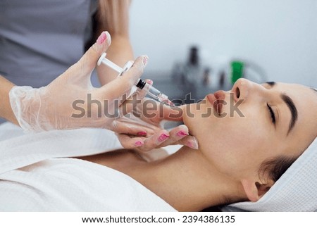 Beauty injections. Female cosmetologist in transparent gloves makes an injection in the lower jaw of her client. Treatment with hyaluronic acid. Introduction of liquid into the skin of woman. Royalty-Free Stock Photo #2394386135