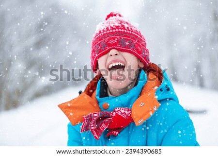 A funny little boy in blue winter clothes is walking during a snowfall. Winter outdoor activities for children. A cute child in a warm hat low above his eyes catches snowflakes with his tongue