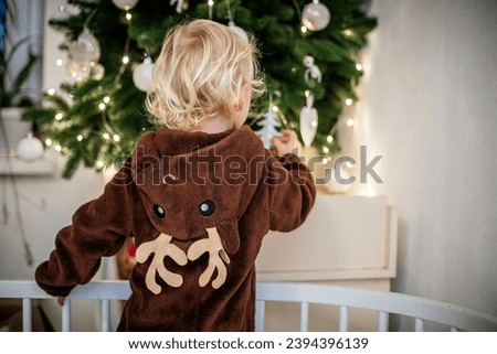 Little girl in reindeer costume decorating Christmas tree while standing in crib Royalty-Free Stock Photo #2394396139
