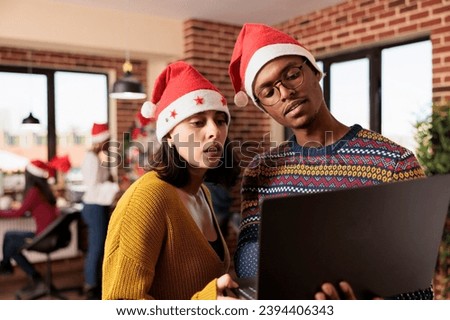 Coworkers in santa hats holding laptop and checking business research report in office during christmas holiday. Diverse colleagues managing workload at xmas eve in workplace