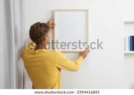 Young man hanging picture frame on white wall indoors, back view Royalty-Free Stock Photo #2394416845