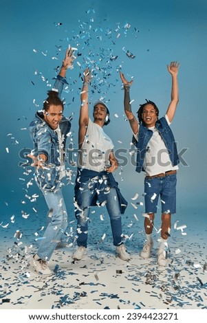 excited multicultural men in trendy denim outfits rejoicing under shiny confetti rain on blue Royalty-Free Stock Photo #2394423271
