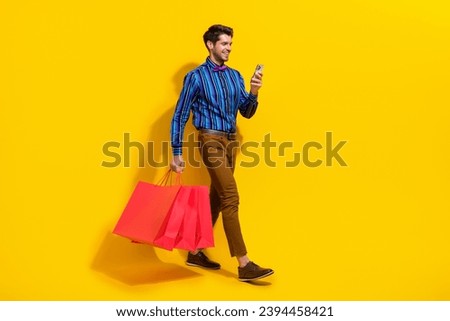 Full size photo of satisfied guy dressed striped shirt go with shopping bags look at smartphone isolated on yellow color background