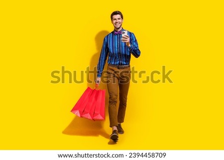 Full size photo of clever guy dressed striped shirt go with shopping bags order clothes on smartphone isolated on yellow color background