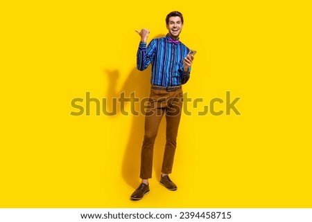 Full size photo of handsome guy dressed striped shirt indicating at sale empty space hold smartphone isolated on yellow color background