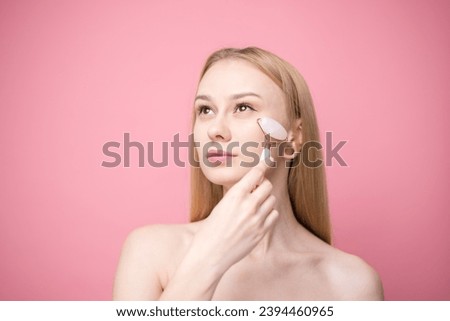 Beautiful young woman giving facial massage with stone roller on pink background. Concept of skin care and aging prevention.