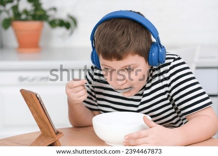 A cute boy of 8 years old watch mobile phone, wear headphones, cartoons and eat at round wooden table in light kitchen, alone at home.