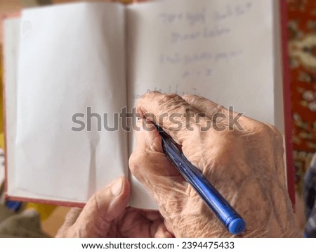 writing, top view selective focused closeup wrinkled hand taking note on a notebook with blue pen. old person writing shopping list or diary on page of a notebook. handwriting concept