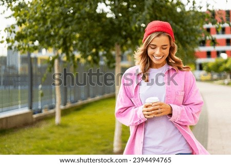 Smiling woman wearing stylish red hat, casual clothes holding cup of coffee looking down, traveling on urban street. Coffee break concept 