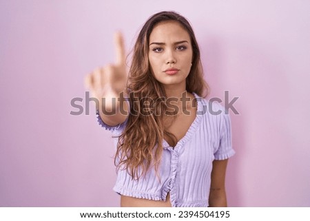 Young hispanic woman standing over pink background pointing with finger up and angry expression, showing no gesture 