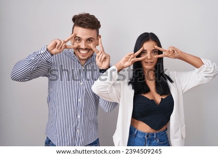 Young hispanic couple standing over white background doing peace symbol with fingers over face, smiling cheerful showing victory 