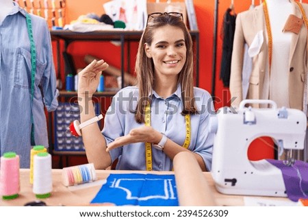 Hispanic young woman dressmaker designer at atelier room gesturing with hands showing big and large size sign, measure symbol. smiling looking at the camera. measuring concept. 