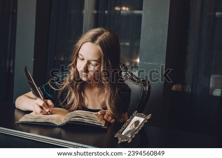 A girl sits in a black office behind an antique steel, reading a book and writing. A little student in her dad's office. A beautiful girl with long hair sits at the table and reads.