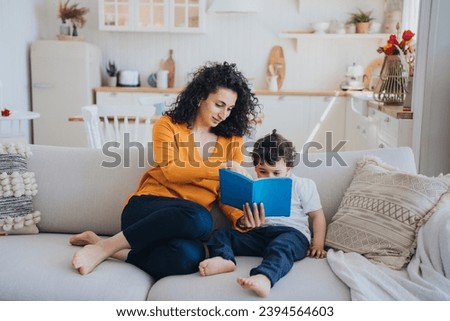 Mother reading book with son sitting on couch at home. Domestic education, childhood. Young mother entertains little boy on weekend. Self isolation, pandemic, stay home. Royalty-Free Stock Photo #2394564603