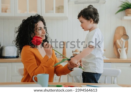 Cute little boy dares to mom beautiful red tulip on mothers day at kitchen. family moments. Children love, motherhood. Young happy woman receiving flower from cute kid home.