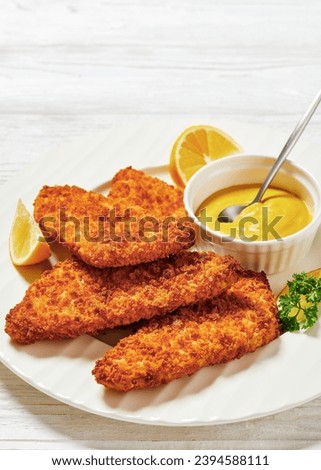 breaded fish fillet baked in oven served with yellow mustard on white plate on white wooden table, vertical view, close-up Royalty-Free Stock Photo #2394588111