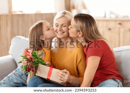 Adoring adult daughter and her young granddaughter kissing their grandmother, joyously celebrating International Womens Day, with the elderly woman receiving gift and bouquet, home interior