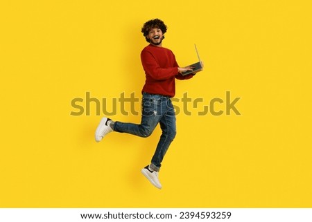 Emotional millennial indian man in casual outfit enjoying great online offer, jumping in the air with laptop computer in his hands, isolated on yellow studio background, copy space Royalty-Free Stock Photo #2394593259